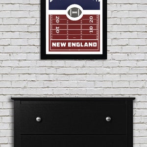May include: A framed print of a football field with a skyline of a city in the background. The field is red and white, and the skyline is white on a navy blue background. The text "NEW ENGLAND" is printed below the field.
