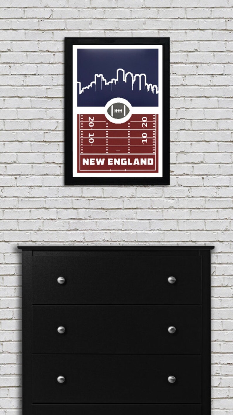 May include: A framed print of a football field with a skyline of a city in the background. The field is red and white, and the skyline is white on a navy blue background. The text "NEW ENGLAND" is printed below the field.