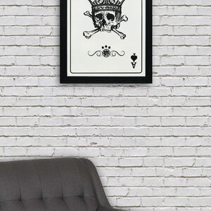 May include: A black and white framed print of an ace of spades playing card with a skull wearing a crown and crossed bones. The card is on a white brick wall background.