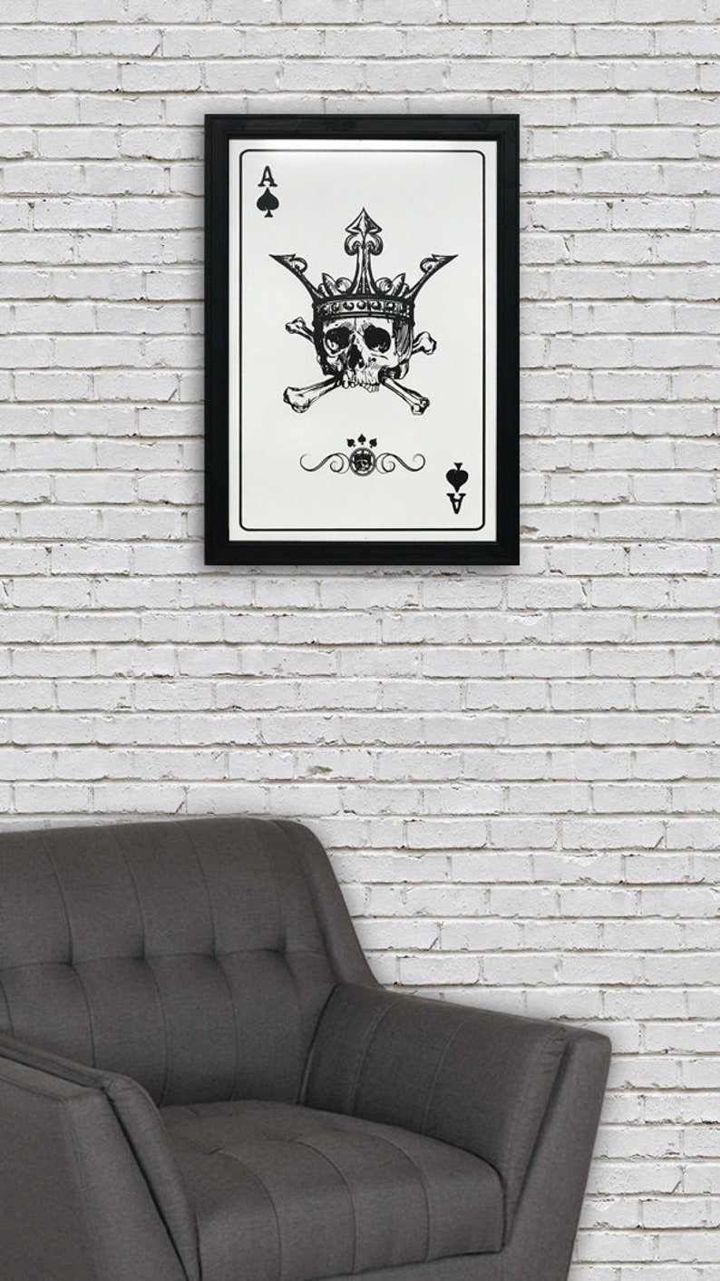 May include: A black and white framed print of an ace of spades playing card with a skull wearing a crown and crossed bones. The card is on a white brick wall background.