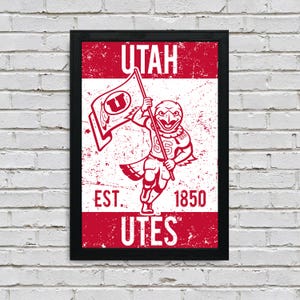 May include: Framed red and white graphic art print featuring the word "UTAH" at the top, a mascot holding a flag with a "U" on it, and the words "EST. 1850 Utes" at the bottom. The artwork has a distressed, vintage look.
