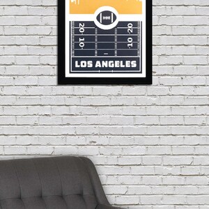 May include: A framed print with a gold background and a black and white silhouette of the Los Angeles skyline. The bottom of the print features a black and white football field with the words "Los Angeles" and "20-10-20".