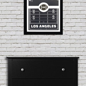 May include: Black and white framed print of a Los Angeles skyline with a football field graphic and the text "LOS ANGELES" underneath. The skyline is depicted in a stylized manner with tall buildings.