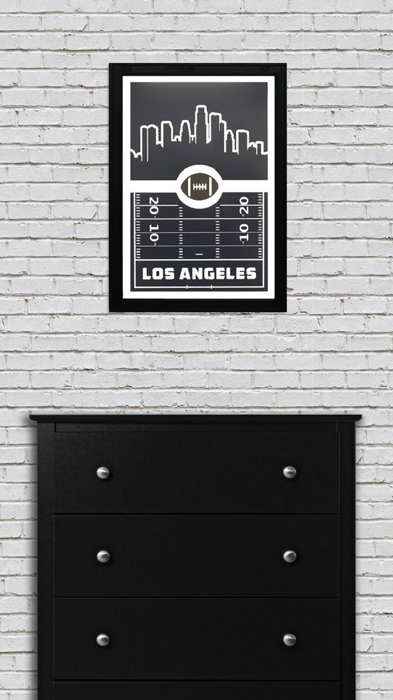 May include: Black and white framed print of a Los Angeles skyline with a football field graphic and the text "LOS ANGELES" underneath. The skyline is depicted in a stylized manner with tall buildings.