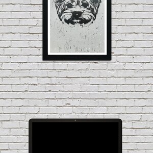 May include: A black and white framed print of a Yorkshire Terrier dog's face. The print is hanging on a white brick wall above a laptop computer.