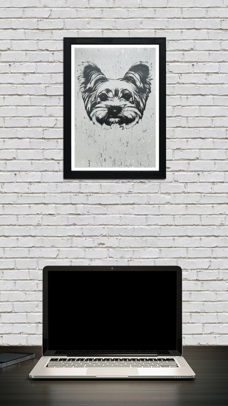 May include: A black and white framed print of a Yorkshire Terrier dog's face. The print is hanging on a white brick wall above a laptop computer.