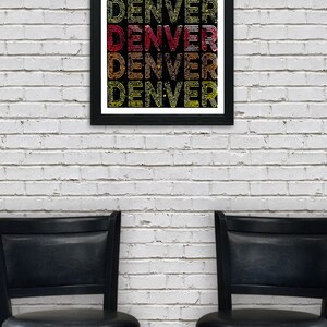 May include: A framed print with the word "DENVER" repeated in a colorful, layered design. The print is in a black frame and is hanging on a white brick wall. Two black bar stools are in the foreground.