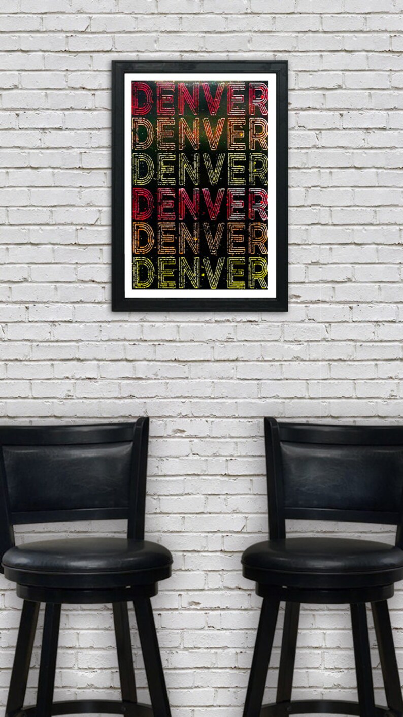 May include: A framed print with the word "DENVER" repeated in a colorful, layered design. The print is in a black frame and is hanging on a white brick wall. Two black bar stools are in the foreground.