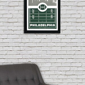 May include: A framed print of a green football field with a white outline of the Philadelphia skyline above it. The text "PHILADELPHIA" is printed below the field.