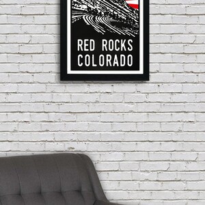 May include: A black and white poster with a red sunburst design. The poster features a silhouette of Red Rocks Amphitheatre in Morrison, Colorado. The text "RED ROCKS COLORADO" is printed in white at the bottom of the poster.