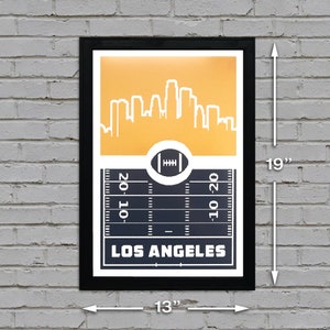 May include: A black framed poster with a gold background featuring a white silhouette of the Los Angeles skyline. The bottom of the poster shows a black and white football field with the text "LOS ANGELES" in white.