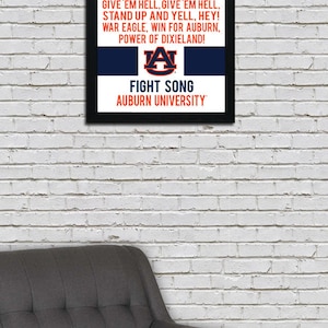 K&ouml;nnte beinhalten: Ein gerahmter Druck mit einem schwarzen Rahmen und einem wei&szlig;en Hintergrund. Der Druck zeigt den Kampfliedtext der Auburn University in blauem und orangefarbenem Text. Der Text enth&auml;lt die W&ouml;rter "War Eagle" und "Fight Song". Das Logo der Auburn University ist in der Mitte des Drucks abgebildet.