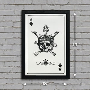 May include: A black framed print of a playing card with a skull wearing a crown and crossed bones. The card is the Ace of Spades. The print measures 13 inches wide by 19 inches tall.