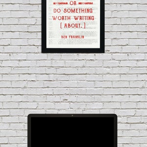 May include: A black framed print with a white background and red text that reads "EITHER WRITE SOMETHING WORTH READING OR DO SOMETHING WORTH WRITING [ABOUT.] BEN FRANKLIN". The print is hanging on a white brick wall. A laptop computer is on a dark surface in front of the print.