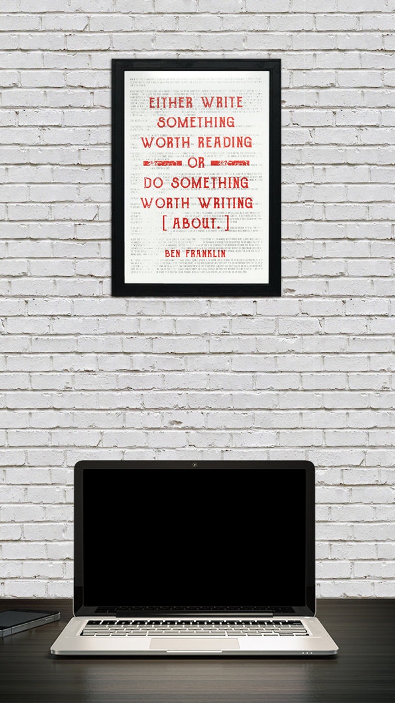 May include: A black framed print with a white background and red text that reads "EITHER WRITE SOMETHING WORTH READING OR DO SOMETHING WORTH WRITING [ABOUT.] BEN FRANKLIN". The print is hanging on a white brick wall. A laptop computer is on a dark surface in front of the print.