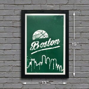 May include: A green and white poster with a basketball and the word "Boston" in a white distressed font. The poster also features a white silhouette of the Boston skyline. The poster is framed in black and measures 13 inches by 19 inches.