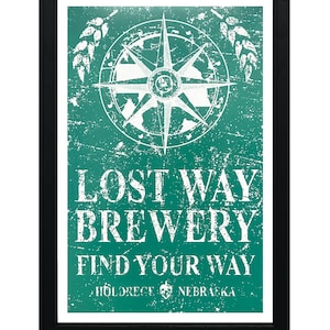 May include: A vintage-style sign with a distressed green background and white text. The sign features a compass with a white star in the center and the words "Lost Way Brewery Find Your Way Holdrege Nebraska".