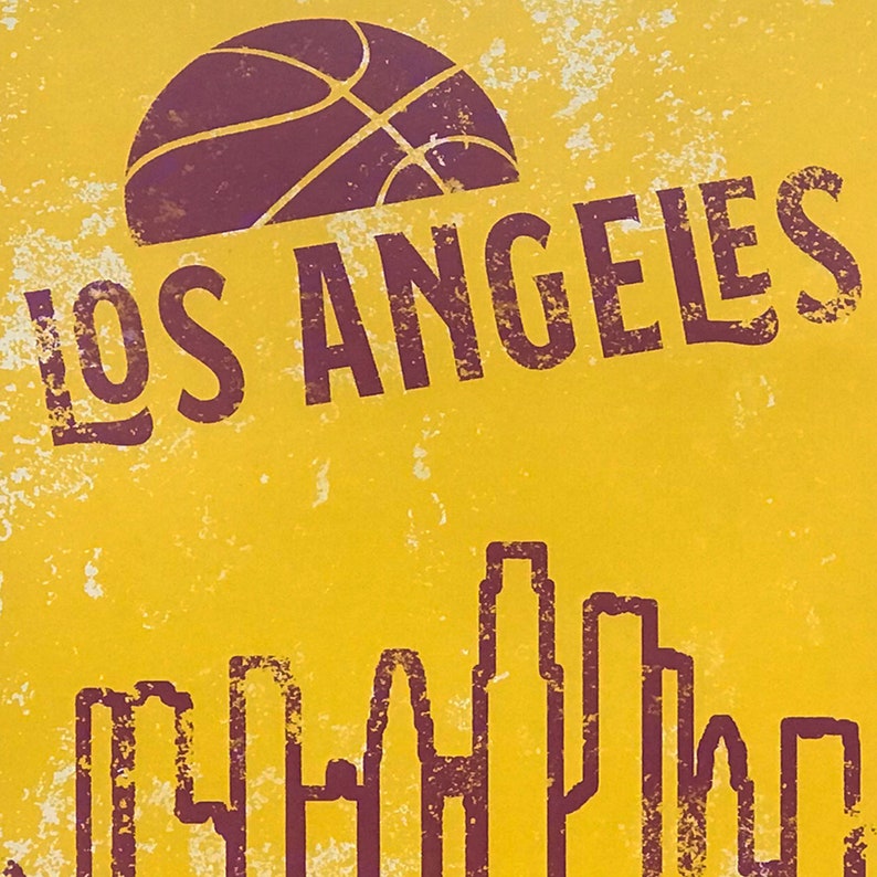 May include: A yellow and purple graphic featuring a basketball and the words "LOS ANGELES" above a skyline silhouette. The design has a distressed, vintage aesthetic, with a textured background.
