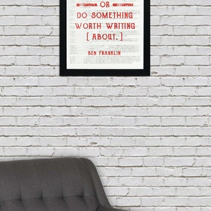 May include: A black framed print with a white background and red text that reads "EITHER WRITE SOMETHING WORTH READING OR DO SOMETHING WORTH WRITING [ABOUT.] BEN FRANKLIN". The print is hanging on a white brick wall above a gray armchair.