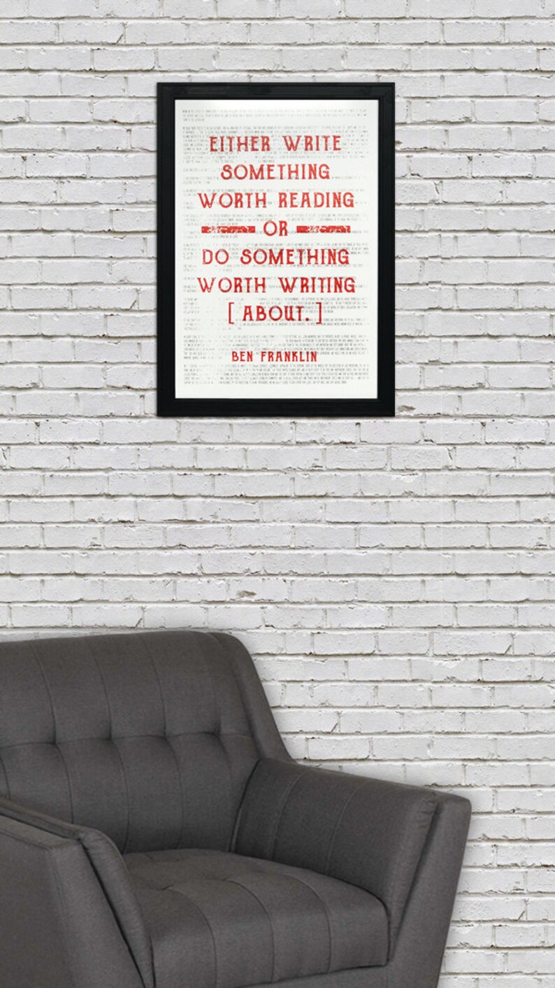 May include: A black framed print with a white background and red text that reads "EITHER WRITE SOMETHING WORTH READING OR DO SOMETHING WORTH WRITING [ABOUT.] BEN FRANKLIN". The print is hanging on a white brick wall above a gray armchair.