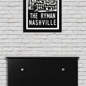 May include: Black and white illustration of The Ryman Auditorium in Nashville, Tennessee. The building is depicted with a red and white sunburst behind it. The text "The Ryman Nashville" is printed below the building.