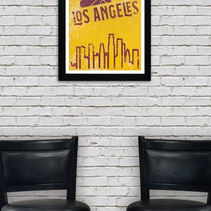 May include: A framed poster with a black border displays a yellow background with a purple basketball and the words "LOS ANGELES". Below the text is a purple skyline graphic. Two black swivel chairs are in front of a white brick wall.