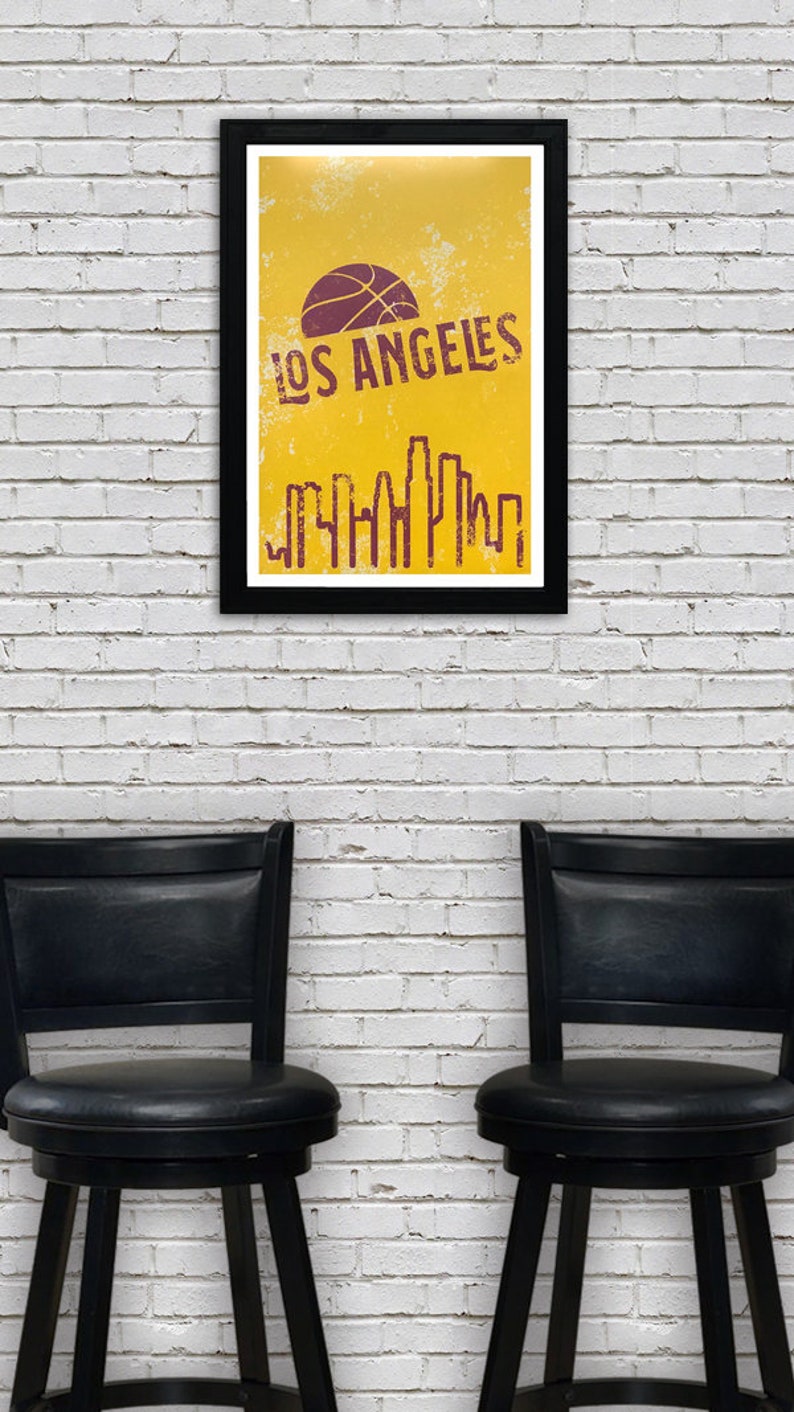 May include: A framed poster with a black border displays a yellow background with a purple basketball and the words "LOS ANGELES". Below the text is a purple skyline graphic. Two black swivel chairs are in front of a white brick wall.