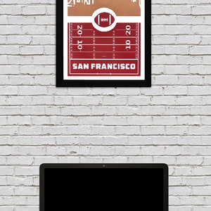 May include: A framed print of a red and white football field with a white outline of the San Francisco skyline above it. The text "SAN FRANCISCO" is printed below the field.