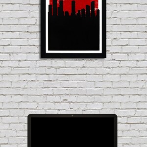 May include: A framed poster with the text "DENVER THE MILE HIGH CITY" in white on a red background, with a black skyline silhouette. Below, a laptop and a smartphone sit on a dark surface against a white brick wall.