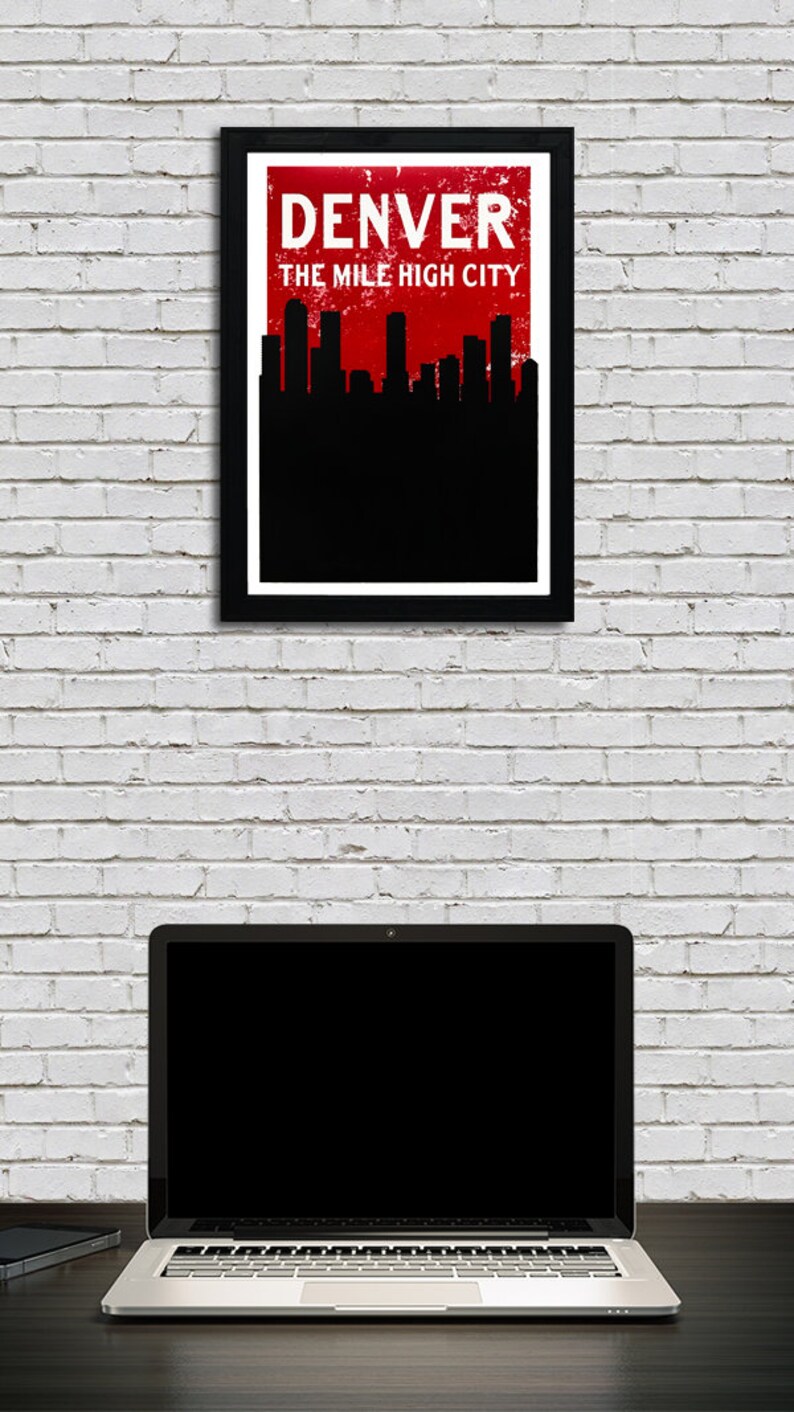 May include: A framed poster with the text "DENVER THE MILE HIGH CITY" in white on a red background, with a black skyline silhouette. Below, a laptop and a smartphone sit on a dark surface against a white brick wall.