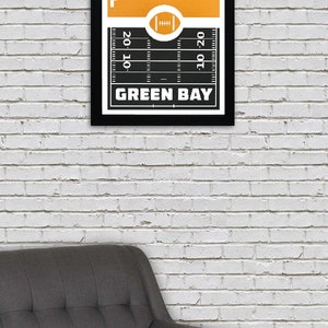 May include: A black and white framed print with a yellow cityscape and a football field. The text "GREEN BAY" is printed below the field.