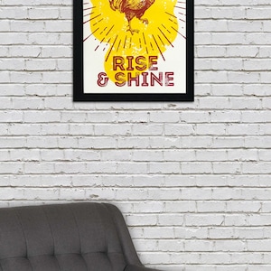 May include: A framed print with a yellow background and a rooster illustration. The rooster is red and yellow and has a red comb. The text "RISE & SHINE" is printed in red below the rooster.