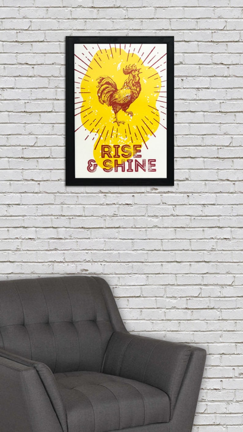 May include: A framed print with a yellow background and a rooster illustration. The rooster is red and yellow and has a red comb. The text "RISE & SHINE" is printed in red below the rooster.