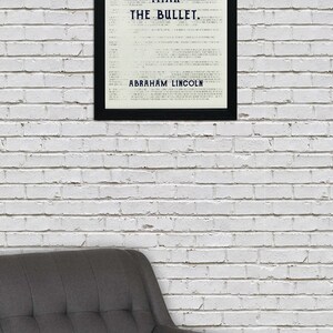 May include: A black framed print with a white background and the text "The ballot is stronger than the bullet. Abraham Lincoln." hanging on a white brick wall above a gray armchair.