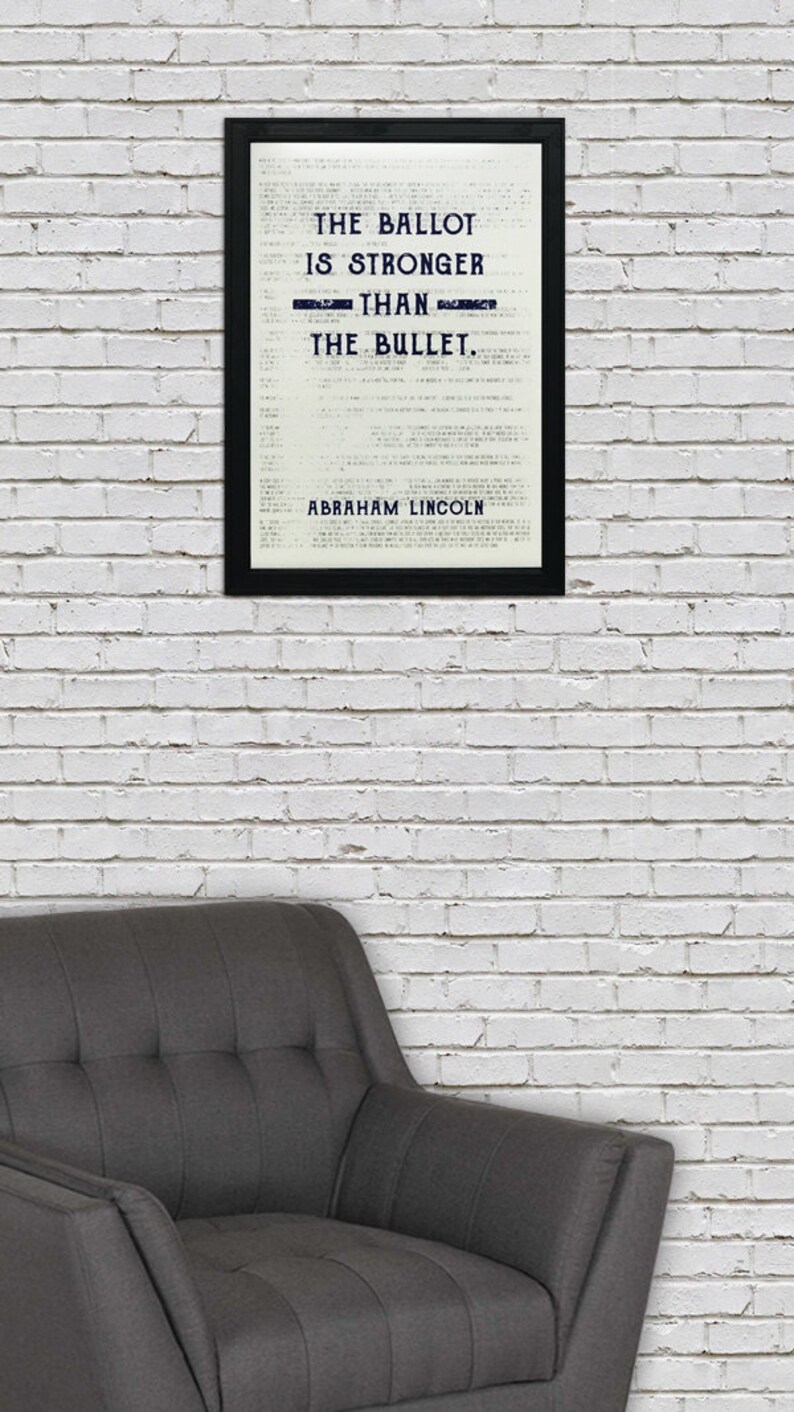 May include: A black framed print with a white background and the text "The ballot is stronger than the bullet. Abraham Lincoln." hanging on a white brick wall above a gray armchair.