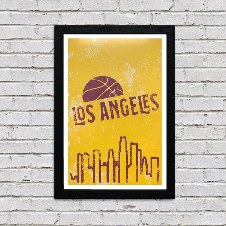 May include: A framed poster with a yellow background and a basketball graphic. The poster features the text "LOS ANGELES" in a distressed font, along with a skyline silhouette. The frame is black, and the poster is mounted on a white brick wall.