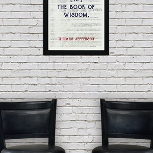 May include: A framed print with the quote "HONESTY IS THE FIRST CHAPTER [IN] THE BOOK OF WISDOM" by Thomas Jefferson. The print is in a black frame, set against a white brick wall. Two black bar stools are in the foreground.