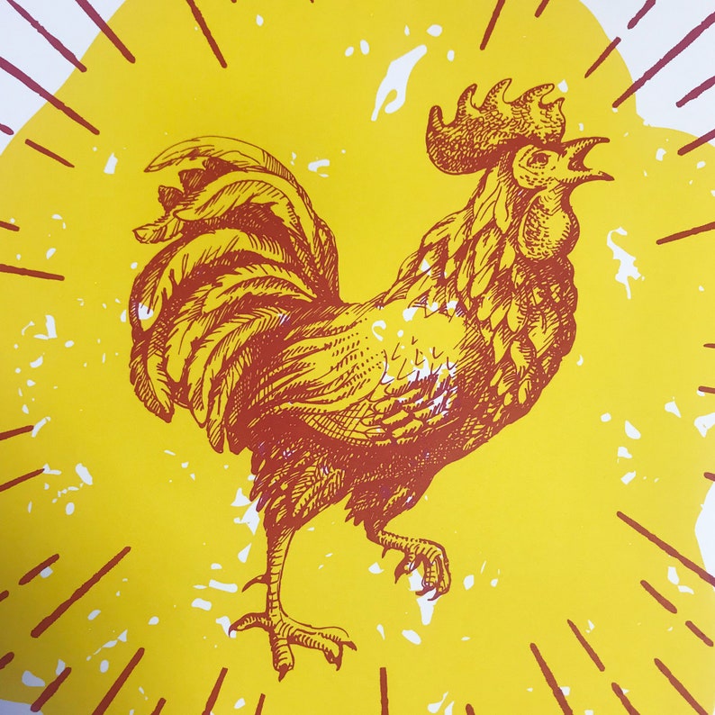 May include: A red rooster illustration with a yellow background. The rooster is facing right and has a large comb and wattles. The rooster is surrounded by a sunburst pattern.