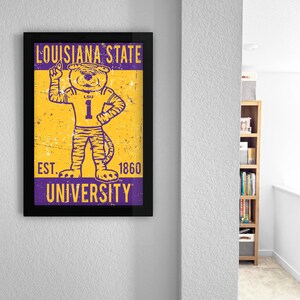 May include: A framed poster with the Louisiana State University tiger mascot in a number 1 jersey. The poster has a distressed, vintage style with the text "LOUISIANA STATE UNIVERSITY" and "EST. 1860". The colours are purple and gold.