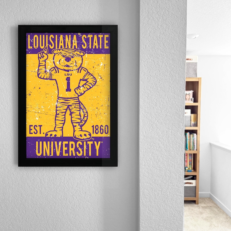 May include: A framed poster with the Louisiana State University tiger mascot in a number 1 jersey. The poster has a distressed, vintage style with the text "LOUISIANA STATE UNIVERSITY" and "EST. 1860". The colours are purple and gold.
