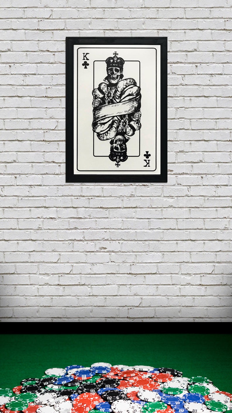 May include: A black and white illustration of the King of Clubs playing card. The King is depicted as a skeleton wearing a crown and holding a scepter. The card is framed in black and hanging on a white brick wall. A pile of poker chips is in the foreground.