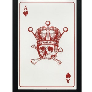 May include: Framed art print featuring a red Ace of Hearts playing card design. The card displays a skull wearing a crown and crossed bones. The artwork is set against a white background and framed in black.