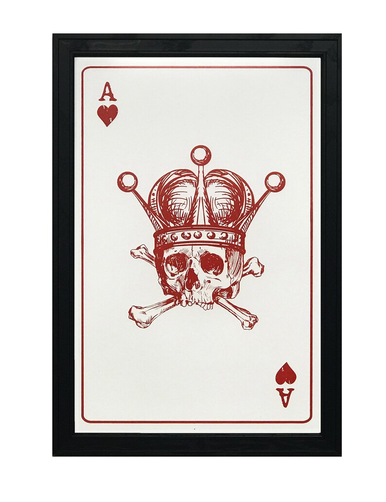 May include: Framed art print featuring a red Ace of Hearts playing card design. The card displays a skull wearing a crown and crossed bones. The artwork is set against a white background and framed in black.
