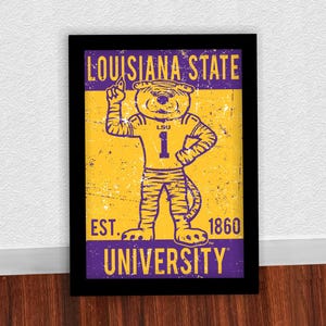 May include: Framed print featuring the Louisiana State University mascot, a tiger wearing a jersey with the number 1. The print has a distressed, vintage look with the words "Louisiana State University" and "Est. 1860". The colour scheme is gold, purple, and black.