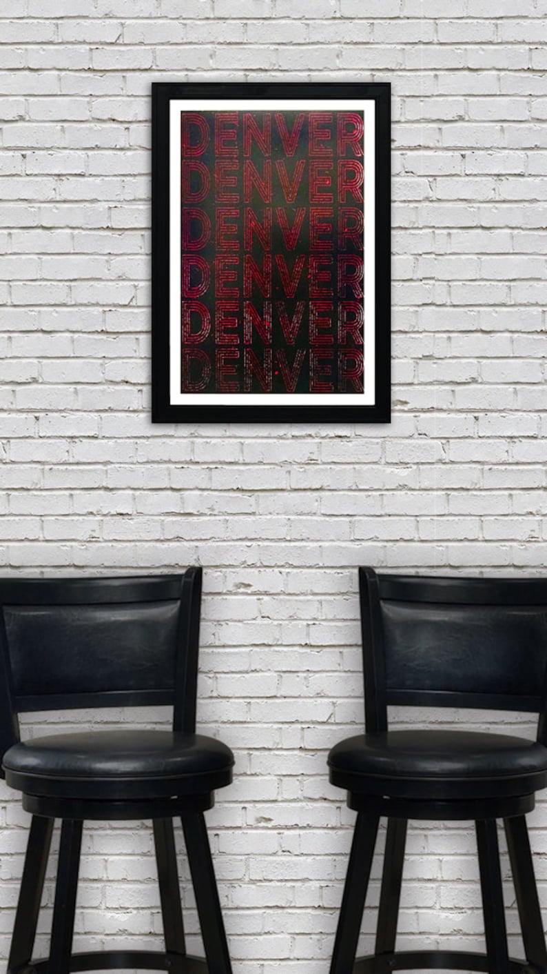May include: A framed print with the word "DENVER" repeated in a red and white striped pattern on a black background. The print is hanging on a white brick wall above two black bar stools.
