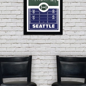 May include: A framed print of a football pitch with a skyline of Seattle, Washington. The skyline includes the Space Needle. The pitch is blue with white yard lines and the words "20-10" and "10-20" on the sides. The words "SEATTLE" are printed below the pitch.