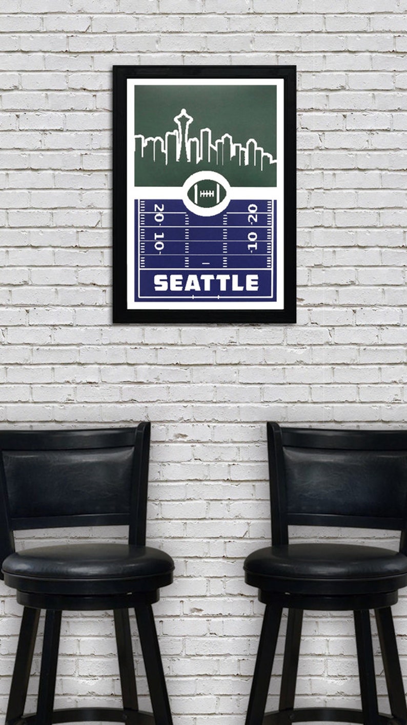 May include: A framed print of a football pitch with a skyline of Seattle, Washington. The skyline includes the Space Needle. The pitch is blue with white yard lines and the words "20-10" and "10-20" on the sides. The words "SEATTLE" are printed below the pitch.