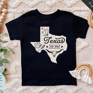 May include: Black t-shirt with a white outline of the state of Texas, a cowboy hat, boots, and a longhorn steer. The text "Texas Est. 1845" is inside the state outline.