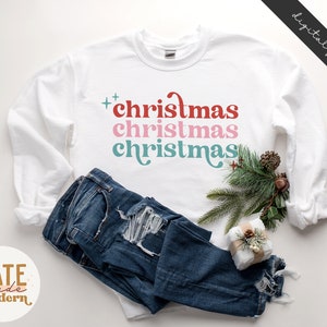 Pode incluir: Camisola branca de manga comprida com o texto "christmas christmas christmas" em vermelho, rosa e azul.