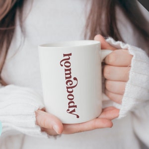 May include: White ceramic mug with the text "homebody" written vertically in a maroon color.